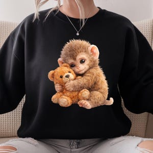 May include: Black sweatshirt featuring a detailed illustration of a brown monkey hugging a brown teddy bear. The monkey has large, expressive eyes and is rendered in a soft, realistic style. The teddy bear is also detailed, with visible stitching and a classic design. The sweatshirt is a solid black color.