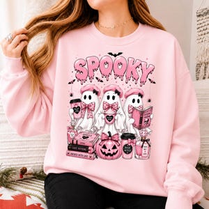 May include: A light pink crewneck sweatshirt featuring a graphic print of three ghosts wearing pink berets and bows. The design includes the text SPOOKY, a pink pumpkin, stacked books labeled SPELLS and LOVE POTIONS, and perfume bottles.