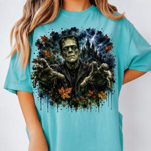May include: A light blue t-shirt featuring a graphic print of a green monster with outstretched hands, a dark castle, lightning, and scattered orange and brown autumn leaves. The design has a watercolor-style ink splatter effect around the edges.