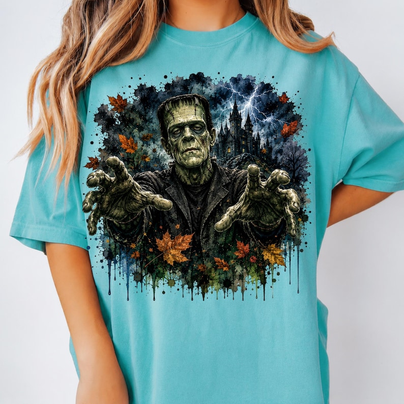 May include: A light blue t-shirt featuring a graphic print of a green monster with outstretched hands, a dark castle, lightning, and scattered orange and brown autumn leaves. The design has a watercolor-style ink splatter effect around the edges.