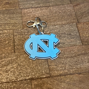 Op de afbeelding: Een lichtblauwe sleutelhanger met het in elkaar grijpende "NC"-logo van de Universiteit van North Carolina. De sleutelhanger heeft een zilverkleurige sluiting en ring voor bevestiging. Het logo is wit omlijnd.
