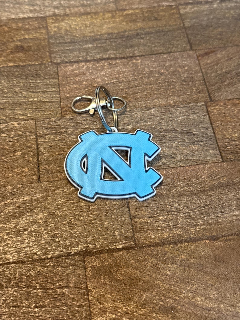 Op de afbeelding: Een lichtblauwe sleutelhanger met het in elkaar grijpende "NC"-logo van de Universiteit van North Carolina. De sleutelhanger heeft een zilverkleurige sluiting en ring voor bevestiging. Het logo is wit omlijnd.