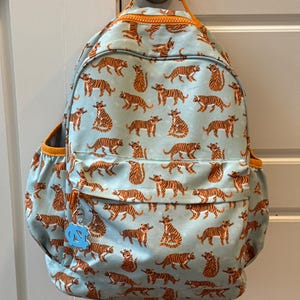 Op de afbeelding: Een lichtblauwe rugzak met een herhaald patroon van oranje tijgers. De rugzak heeft oranje afwerking, een zijvak en een kleine bedel. De rugzak hangt aan een deur.