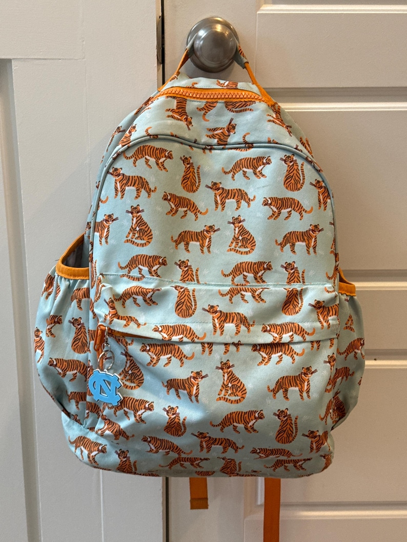 Op de afbeelding: Een lichtblauwe rugzak met een herhaald patroon van oranje tijgers. De rugzak heeft oranje afwerking, een zijvak en een kleine bedel. De rugzak hangt aan een deur.