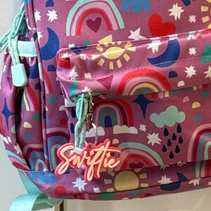 May include: A purple backpack with a colorful rainbow, sun, and star pattern. The backpack has a front pocket and a light blue zipper pull. A keychain with the word "Swiftie" hangs from the zipper.
