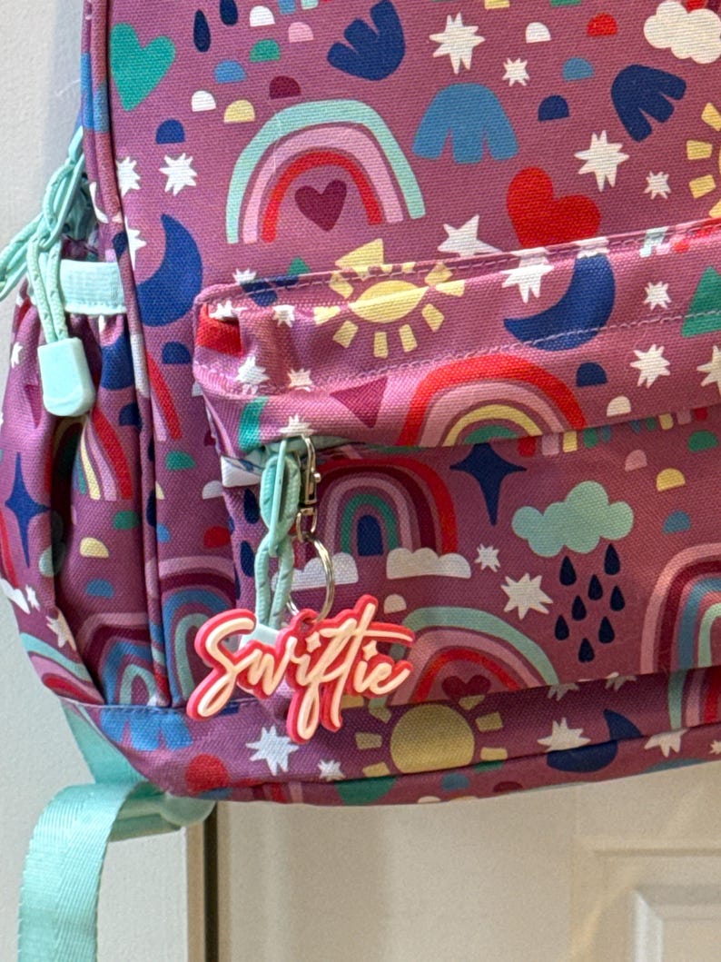 May include: A purple backpack with a colorful rainbow, sun, and star pattern. The backpack has a front pocket and a light blue zipper pull. A keychain with the word "Swiftie" hangs from the zipper.