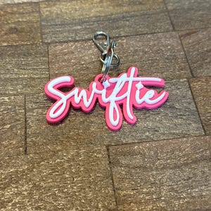 May include: A pink and white keychain with the word "Swiftie" in a cursive font. The keychain has a silver clasp and ring for attaching to keys or a bag. The background is a brown, wood-like surface.