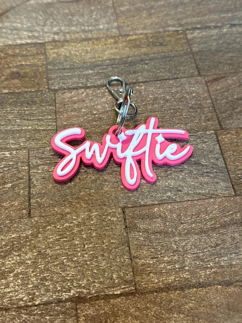 May include: A pink and white keychain with the word "Swiftie" in a cursive font. The keychain has a silver clasp and ring for attaching to keys or a bag. The background is a brown, wood-like surface.