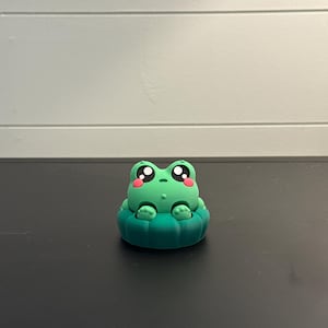 May include: A small, green frog figurine with large, round eyes and rosy cheeks. The frog is sitting in a lily pad-shaped base. The figurine is made of a solid material and has a smooth finish.