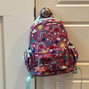 May include: A purple backpack with a colorful rainbow and sun pattern. The backpack has a front pocket and a light blue handle and straps. The word "Swiftie" is written on a tag attached to the zipper.
