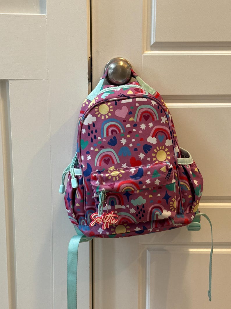 May include: A purple backpack with a colorful rainbow and sun pattern. The backpack has a front pocket and a light blue handle and straps. The word "Swiftie" is written on a tag attached to the zipper.
