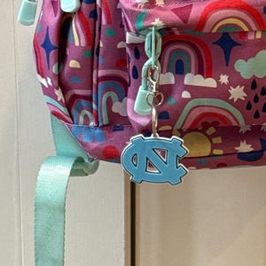Op de afbeelding: Een paarse rugzak met een regenboog- en zonnepatroon, met een lichtblauwe ritstrekker en riem. Een lichtblauwe sleutelhanger met het logo van de Universiteit van North Carolina hangt aan de rits.