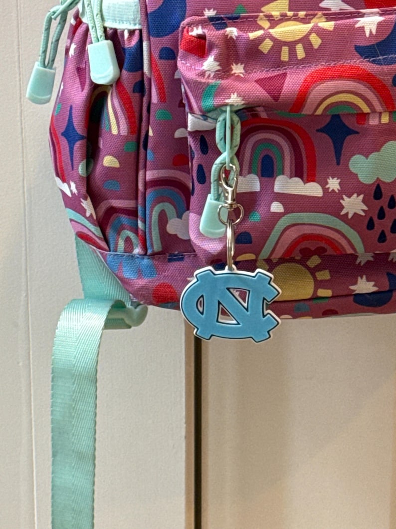 Op de afbeelding: Een paarse rugzak met een regenboog- en zonnepatroon, met een lichtblauwe ritstrekker en riem. Een lichtblauwe sleutelhanger met het logo van de Universiteit van North Carolina hangt aan de rits.