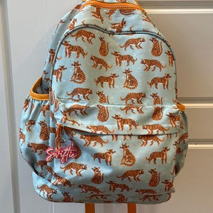 May include: A light blue backpack with an all-over pattern of orange tigers. The backpack has orange trim, a zipper, and side pockets. A pink tag with the name "Swiftie" hangs from the zipper.