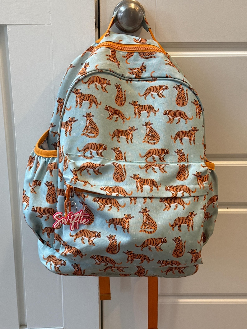 May include: A light blue backpack with an all-over pattern of orange tigers. The backpack has orange trim, a zipper, and side pockets. A pink tag with the name "Swiftie" hangs from the zipper.