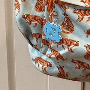 Op de afbeelding: Een lichtblauwe rugzak met een herhaald patroon van oranje en zwarte tijgers. Een lichtblauwe sleutelhanger met een witte omtrek van de letters "NC" hangt aan de rits. De rugzak heeft een oranje rits en een zilveren sleutelring.