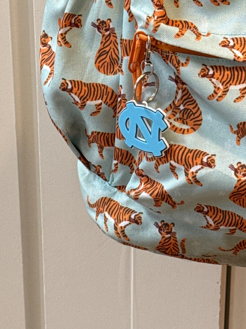 Op de afbeelding: Een lichtblauwe rugzak met een herhaald patroon van oranje en zwarte tijgers. Een lichtblauwe sleutelhanger met een witte omtrek van de letters "NC" hangt aan de rits. De rugzak heeft een oranje rits en een zilveren sleutelring.