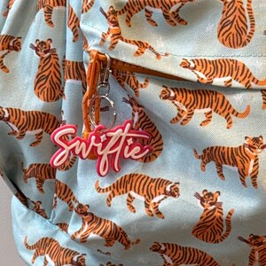 May include: A light blue backpack with a repeating pattern of orange and black tigers. The backpack has an orange zipper pull and a pink and white keychain that says "Swiftie".