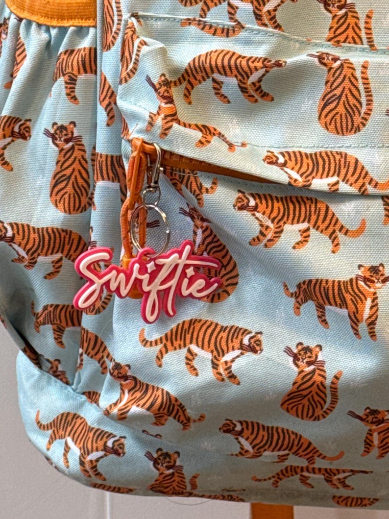 May include: A light blue backpack with a repeating pattern of orange and black tigers. The backpack has an orange zipper pull and a pink and white keychain that says "Swiftie".