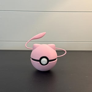 May include: A pink, spherical object with a black band and a white button. It has two curved pink arms and a pink, leaf-shaped extension. The object is on a black surface.