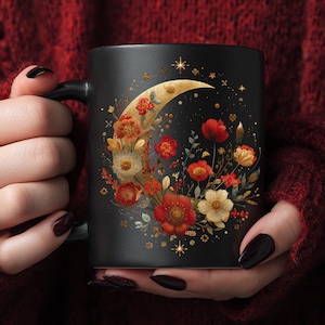 May include: Black ceramic mug featuring a crescent moon design filled with red, orange, and cream-colored flowers and gold accents. The mug has a black handle and a floral and celestial theme.