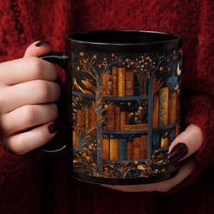 Puede incluir: Una taza de cer&aacute;mica negra con una ilustraci&oacute;n detallada de una estanter&iacute;a llena de libros antiguos. El dise&ntilde;o incorpora detalles dorados y azules, con una luna creciente y ramas de &aacute;rboles. La taza es sostenida por una persona que lleva un su&eacute;ter rojo.