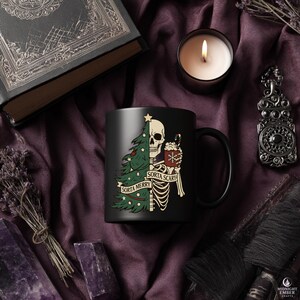 May include: Black ceramic mug featuring a skeleton design with a Christmas tree and the text "Sorta Merry Sorta Scary." The mug also has a candy cane and a cup of hot cocoa. The mug is surrounded by a candle, a book, and other decorative items.