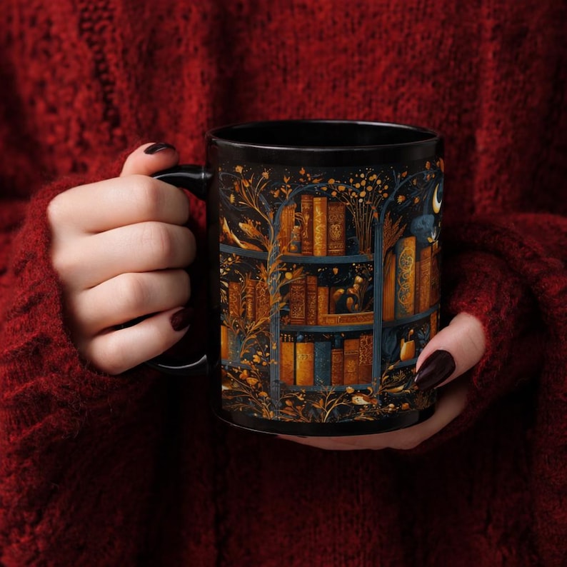 Puede incluir: Una taza de cer&aacute;mica negra con una ilustraci&oacute;n detallada de una estanter&iacute;a llena de libros antiguos. El dise&ntilde;o incorpora detalles dorados y azules, con una luna creciente y ramas de &aacute;rboles. La taza es sostenida por una persona que lleva un su&eacute;ter rojo.