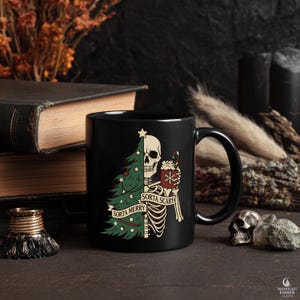 May include: Black ceramic mug with a humorous Christmas design. One side features a green Christmas tree, the other a skeleton holding a mug of cocoa. The text reads "Sorta Merry" and "Sorta Scary".