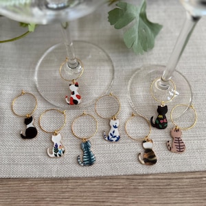 May include: A collection of cat-shaped wine charms. Each charm is a different color and pattern, including black, white, calico, and blue. The charms hang from gold-colored hoops.