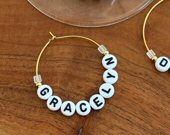 May include: Gold hoop earrings with white letter beads spelling out "GRACE" and "CAMDEN". The earrings are displayed on a wooden surface. The letters are black and the beads are white. The earrings are a personalized jewelry item.