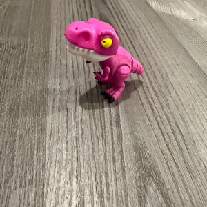 Puede incluir: Un juguete de dinosaurio rosa con mandíbula blanca y ojos amarillos. El dinosaurio está sobre una superficie de madera. El juguete tiene extremidades articuladas y un diseño detallado.
