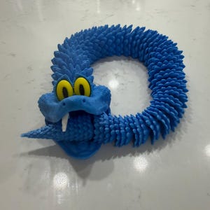 Può includere: Un giocattolo blu a forma di serpente con occhi gialli e una zanna bianca. Il giocattolo è realizzato in un materiale strutturato ed è avvolto a forma di anello. La testa del serpente è nella parte anteriore, con la bocca leggermente aperta.