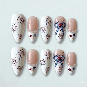 May include: A set of ten artificial nails with a white base, featuring red, white, and blue fireworks designs. Some nails have a nude-colored tip with a pearl and star accent, while others showcase a blue bow.