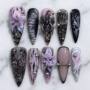 Op de afbeelding: Een verzameling van tien lange, puntige kunstnagels met ingewikkelde ontwerpen. De nagels hebben verschillende stijlen, waaronder bloemaccenten, kanten patronen en metalen versieringen. Kleuren zijn roze, zwart, grijs en zilver, wat een gedetailleerde en artistieke weergave creëert.