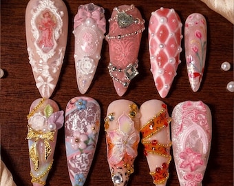 Puede incluir: Un juego de diez uñas postizas en forma de almendra con intrincados diseños en 3D. Los adornos incluyen marcos barrocos blancos, lazos rosas, perlas, cadenas plateadas, pedrería, motivos florales, cintas doradas y patrones texturizados en tonos rosa y crema.