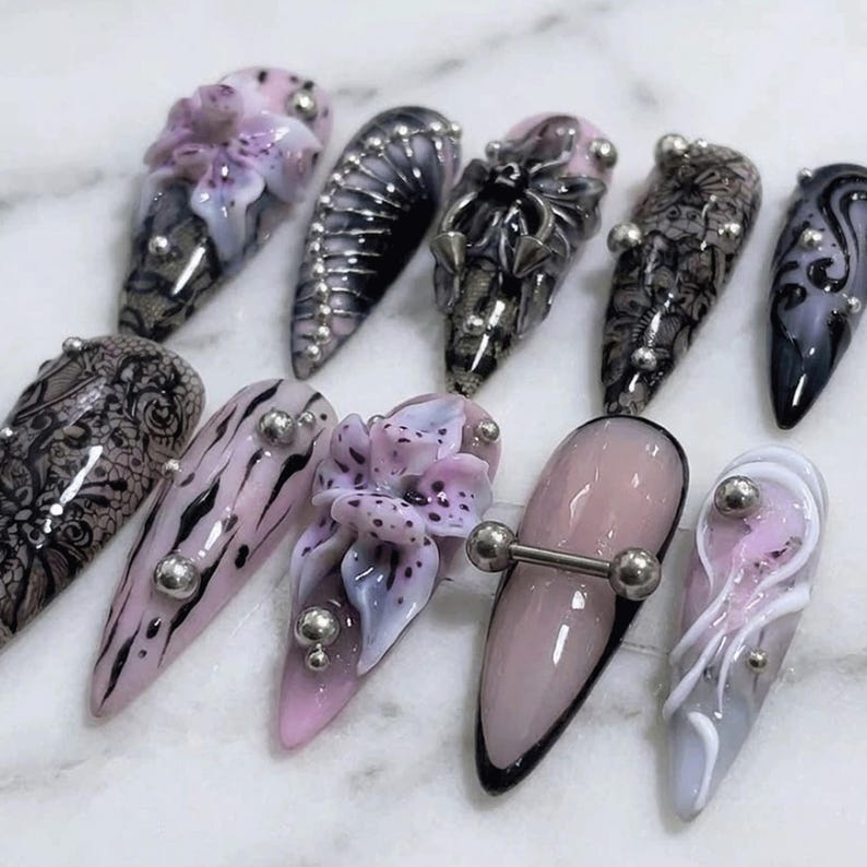 May include: A selection of ten long, pointed artificial nails with diverse designs. The nails showcase pink, black, grey, and white hues, adorned with floral patterns, lace, and silver studs. Some nails incorporate a piercing-style bar.