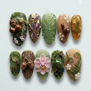 May include: A collection of ten intricately designed artificial nails. Each nail features a unique nature-inspired design, including mushrooms, flowers, insects, and foliage. The color palette includes greens, browns, golds, and pinks, creating a detailed and artistic display.
