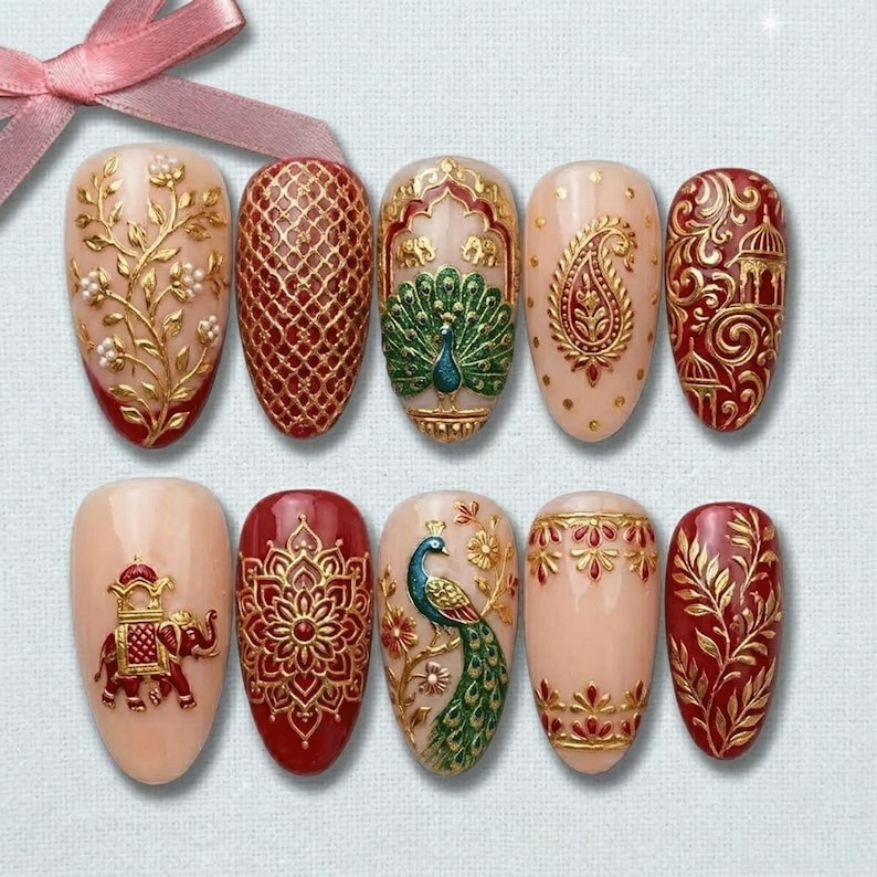 May include: A set of ten artificial nails with intricate designs. The nails feature a variety of patterns, including gold floral, peacock, and paisley motifs, set against a nude and red base. The designs are detailed and ornate, with a 3D effect.