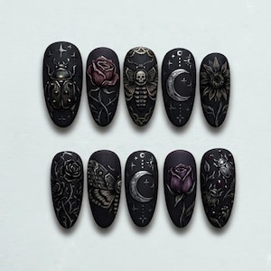 May include: A set of ten black, almond-shaped artificial nails. Each nail features a different design, including a beetle, rose, moth with a skull, crescent moon, and sunflower. The designs are in shades of gray, white, and purple.