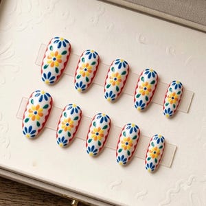 Peut inclure: Un ensemble de dix faux ongles en forme d'amande avec une base blanche et un motif floral peint à la main. Chaque ongle présente une fleur jaune centrale entourée de pétales bleus et de feuilles vertes, avec une bordure rouge festonnée.