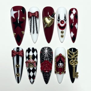 May include: A set of ten long, pointed artificial nails with various designs. The nails feature a color palette of red, black, white, and gold. Designs include stars, bows, harlequin patterns, a clown face, a spiderweb, and a key.