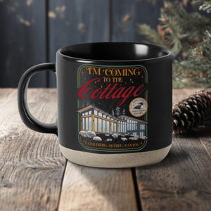 May include: Black ceramic mug with a cream-colored base. The mug features a graphic with the text "I'M COMING TO THE Cottage" and an illustration of a modern cabin. The text "Lanaudiere, Quebec, Canada" is also included.