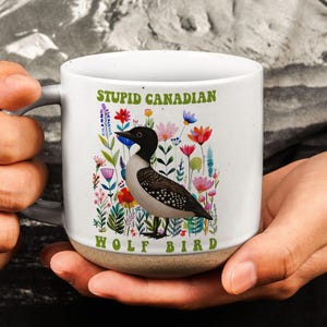 Può includere: Tazza in ceramica bianca con un'illustrazione di un uccello nero, bianco e marrone circondato da fiori colorati. La tazza ha il testo "STUPID CANADIAN WOLF BIRD" in verde. La tazza è tenuta da una persona.