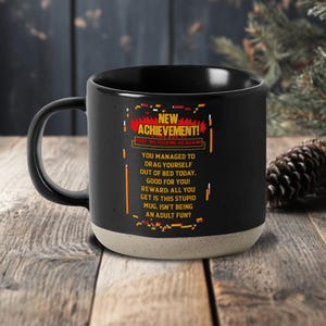 May include: Black ceramic mug with a beige base, featuring the text "NEW ACHIEVEMENT!" and a humorous message about adulting. The mug has a black handle and is set on a wooden surface.