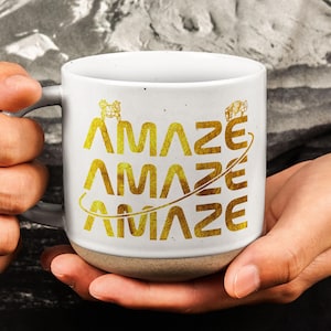 Könnte beinhalten: Weißer Keramikbecher mit grauem Henkel und beigefarbenem Boden. Der Becher zeigt das Wort "AMAZE" in Gold, dreimal wiederholt, mit einer goldenen Linie und Illustration. Der Becher wird von einer Person gehalten.