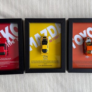 May include: Three framed toy car displays. The first has a red car on a red background with white text. The second has a yellow car on a yellow background. The third has an orange car on an orange background.
