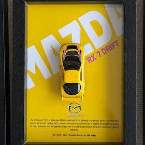 Puede incluir: Impresión enmarcada con un coche de juguete Mazda RX-7 Drift amarillo. El coche tiene detalles en rosa y se encuentra sobre un fondo amarillo con el texto "MAZDA RX-7 DRIFT". El marco es negro.