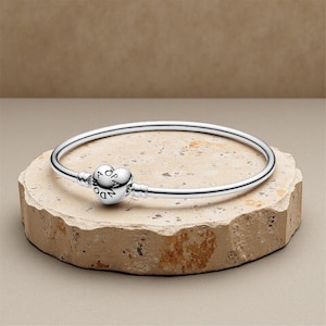 May include: A silver bangle bracelet with a heart-shaped clasp. The clasp features the word "Pandora" and is centered on a stone surface. The bracelet is a simple, elegant design, perfect for everyday wear. The bracelet is approximately 7 inches in diameter.
