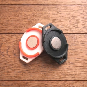 May include: Two circular, 3D-printed objects with a magnetic center. One is white with orange accents, the other is black. Both have a loop for attachment. The objects are displayed on a wooden surface.